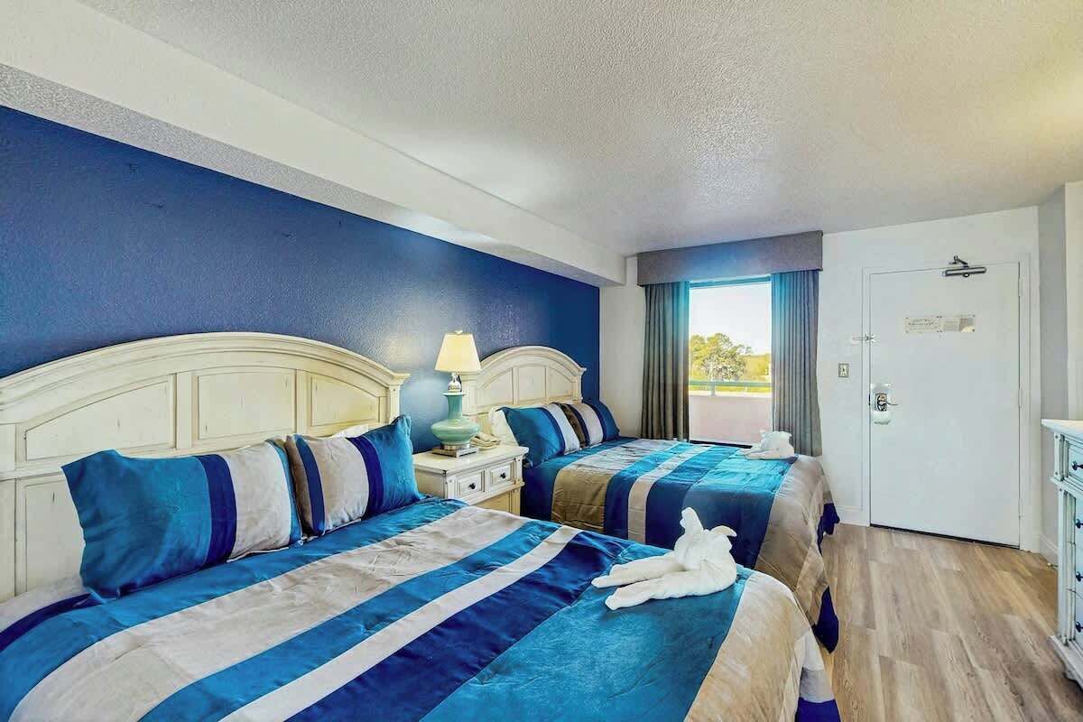 A hotel room with two beds, a nightstand with a lamp, a door, and a view of the outdoors.