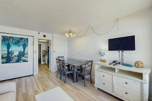 A dining area with a table and chairs, a television on a stand, and a painting on the wall. - Sandcastle South 615 (Myrtle Beach)