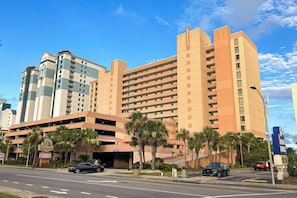 A multi-story hotel with a parking garage, palm trees, and a clear sky. - Sandcastle South 615 (Myrtle Beach)