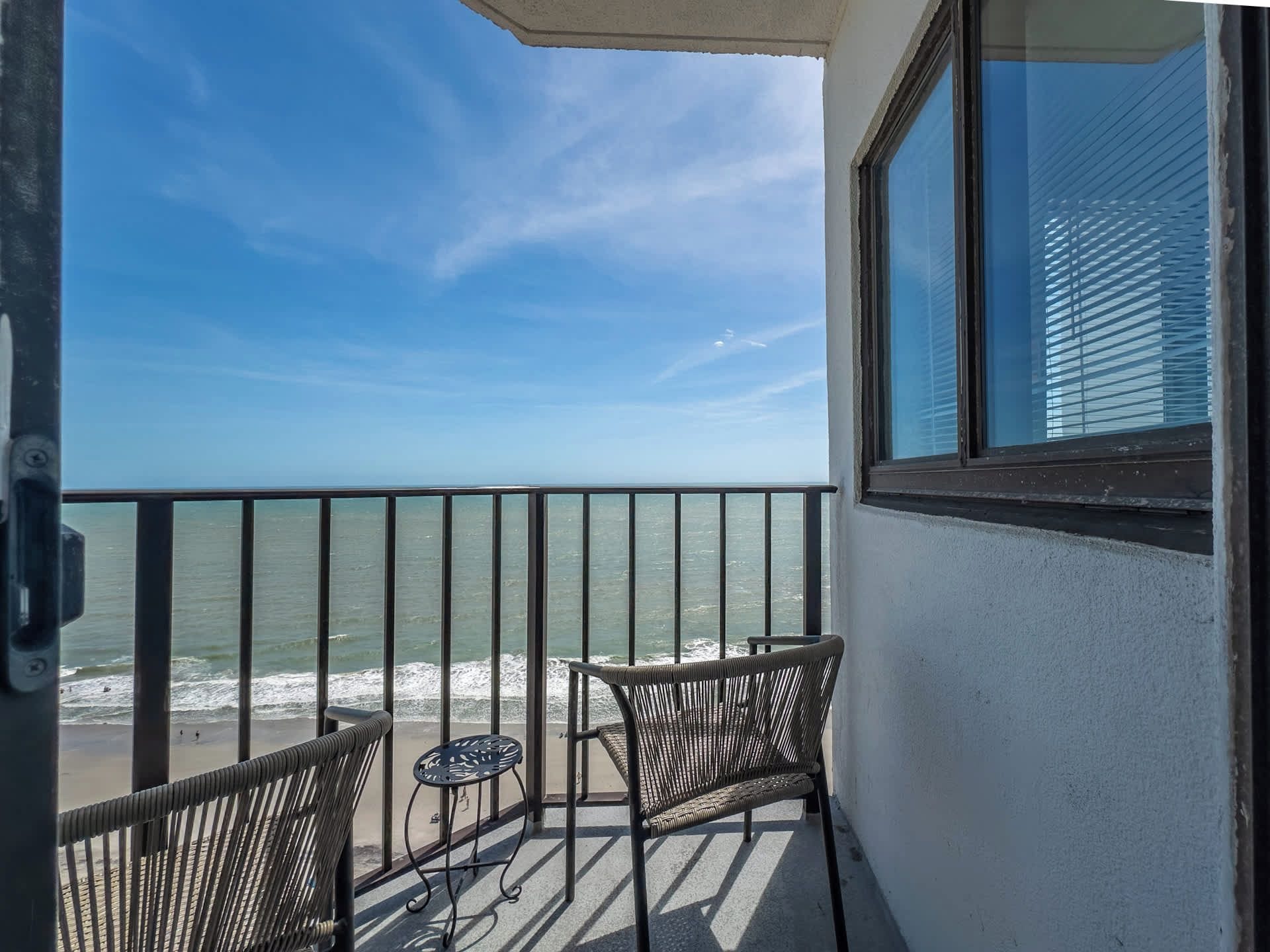 A balcony with two wicker chairs and a small table, overlooking a beach with waves.