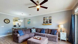 A living room with a sectional sofa, ottoman, ceiling fan, dining area, and artwork on the walls. - Wedgewood Unit 423 Condo (North Myrtle Beach)