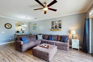 Ein Wohnzimmer mit Ecksofa, Hocker, Deckenventilator, Essbereich und Bildern an den Wänden.