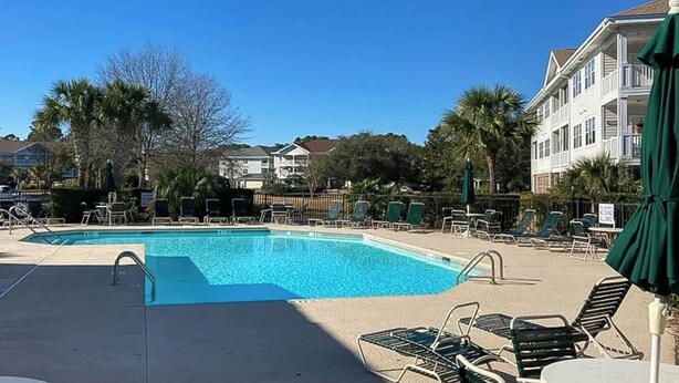 A swimming pool with lounge chairs and umbrellas, surrounded by buildings and palm trees. - Wedgewood Unit 423 Condo (North Myrtle Beach)