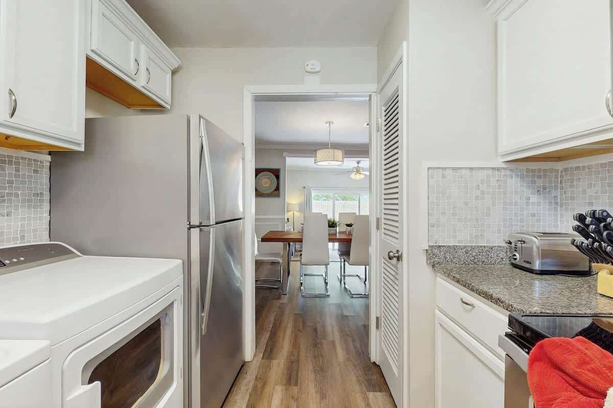 A compact kitchen with white cabinetry, a stainless steel refrigerator, a toaster, and a wooden dining table with chairs.