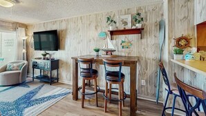 A room with a wooden wall, a flat-screen TV, a wooden dining table with chairs, a small shelf with decorative items, and a large window. - Ocean Pier IV Unit - 332 (North Myrtle Beach)