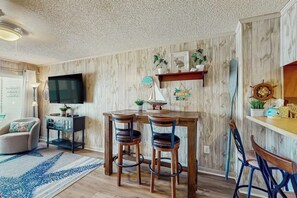 A room with a wooden wall, a flat-screen TV, a wooden dining table with chairs, a small shelf with decorative items, and a large window. - Ocean Pier IV Unit - 332 (North Myrtle Beach)