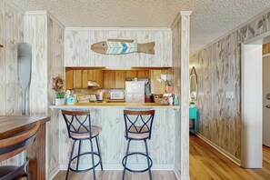 A kitchen with wooden paneling, a fish-shaped decoration, a bar area with stools, and a refrigerator. - Ocean Pier IV Unit - 332 (North Myrtle Beach)