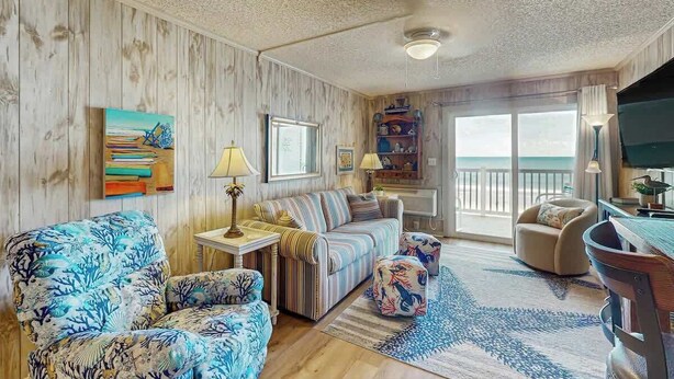 A room with a striped sofa, a patterned armchair, a small table with a lamp, and a view of the outdoors through a glass door. - Ocean Pier IV Unit - 332 (North Myrtle Beach)