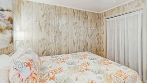 A bedroom with a bed, a nightstand, a wall with a floral pattern, and a window with curtains. - Ocean Pier IV Unit - 332 (North Myrtle Beach)