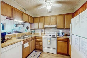 A kitchen with wooden cabinets, a white stove, a ceiling fan, and a refrigerator with papers on it. - Ocean Pier IV Unit - 332 (North Myrtle Beach)