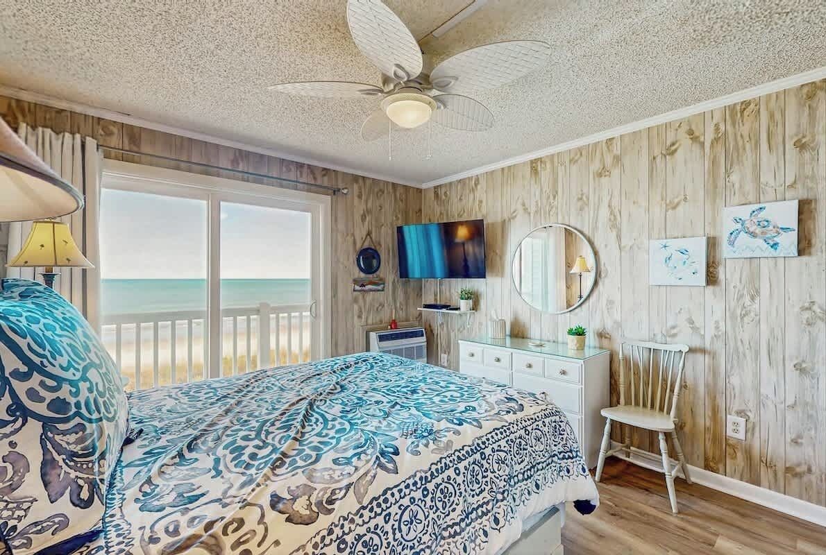 Une chambre à coucher comprenant un lit, une commode, une chaise, un ventilateur de plafond, un téléviseur et une vue sur l’océan.