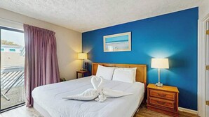 A hotel room with a bed, two bedside lamps, a window with curtains, a painting on the wall, and a wooden nightstand. - The Arbor Unit 303 Condo (Myrtle Beach)