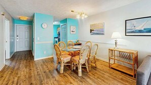 A dining area with a round table and chairs, a wall clock, and a framed picture. - The Arbor Unit 303 Condo (Myrtle Beach)