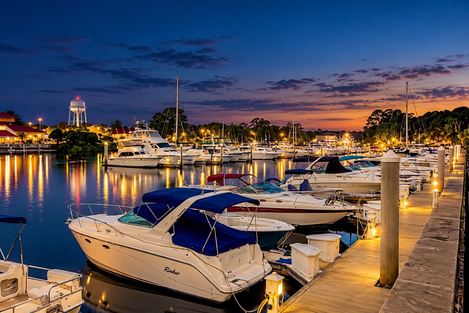A marina at dusk with boats docked, a water tower in the background, and a docked boat with a name 'Rinker' visible.