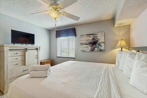 A hotel room with a bed, a television, a ceiling fan, a window with blinds, a painting on the wall, and a bedside lamp. - Yacht Club Villas 1-501 (North Myrtle Beach)