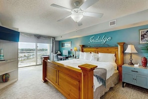 A bedroom with a bed, a ceiling fan, a nightstand, a lamp, and a view of the outdoors. - Yacht Club Villas 1-501 (North Myrtle Beach)