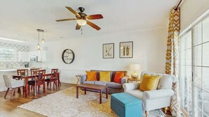 Ein Wohnzimmer mit einer Couch, einem Couchtisch und Sesseln, einem Deckenventilator und einem Essbereich mit Tisch und Stühlen.