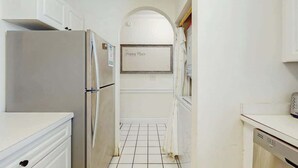 Eine schmale Küche mit weißen Schränken, einem Edelstahl-Kühlschrank, einem Fliesenboden und einem an der Wand befestigten Schild, auf dem 'Happy Place' steht.