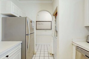 Eine schmale Küche mit weißen Schränken, einem Edelstahl-Kühlschrank, einem Fliesenboden und einem an der Wand befestigten Schild, auf dem 'Happy Place' steht.