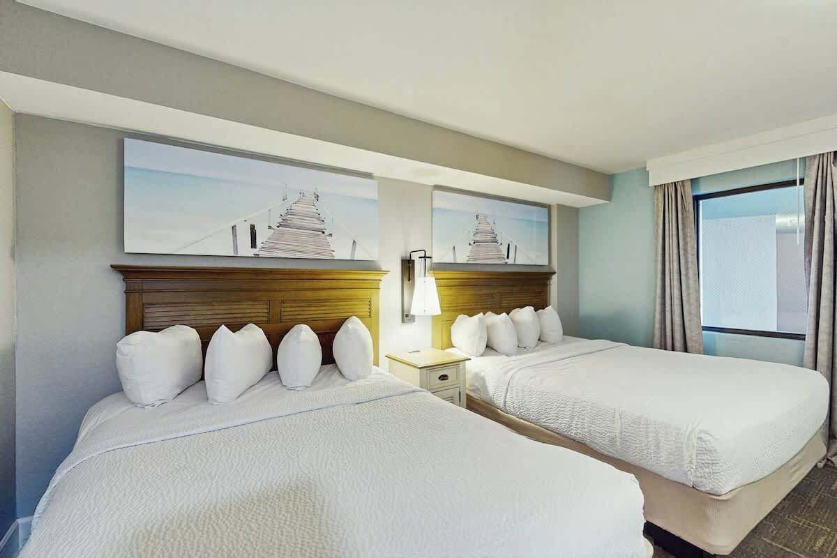 A hotel room with two beds, a nightstand, a lamp, and a window with curtains.