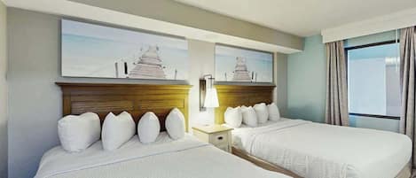 Quarto de hotel com duas camas, uma mesa de cabeceira, uma luminária e uma janela com cortinas.