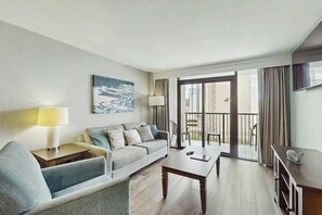 A modern hotel room with a sofa, a coffee table, a lamp, and a view of the city through large windows. - Compass Cove Unit 824 (Myrtle Beach)