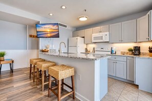 A modern kitchen with a large island, bar stools, a flat-screen TV, and a breakfast bar. - Renaissance Luxe | Two Sixteen C (Myrtle Beach)