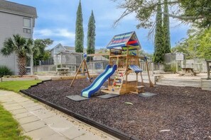 A playground with a slide and climbing structures, surrounded by a fence and trees. - Arcadian Dunes 04-213 (Myrtle Beach)