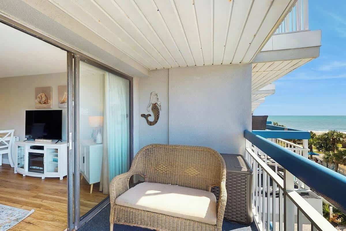 Un balcon avec un fauteuil en osier, une porte vitrée donnant sur une pièce équipée d’un téléviseur et de meubles blancs, et une vue sur la mer.