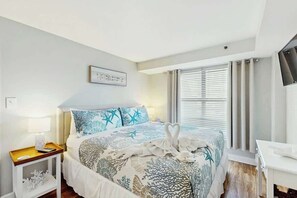 A hotel room with a bed, a bedside table with a lamp, a TV on the wall, and a window with curtains. - Forest Dunes 401 | King Bed, Ocean View & Pools (Myrtle Beach)