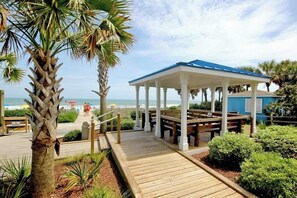 A wooden boardwalk leading to a covered pavilion with benches, palm trees, and a view of the beach. - Forest Dunes 401 | King Bed, Ocean View & Pools (Myrtle Beach)
