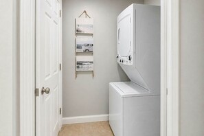 A laundry room with a washing machine, a drying rack, and a wall-mounted shelf with pictures. - Forest Dunes 401 | King Bed, Ocean View & Pools (Myrtle Beach)