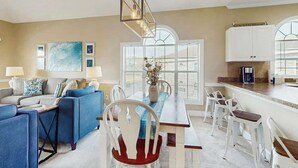 Uma sala de estar moderna com sofá azul, cadeiras brancas, uma mesa de jantar e uma área de cozinha com armários brancos e uma cafeteira.