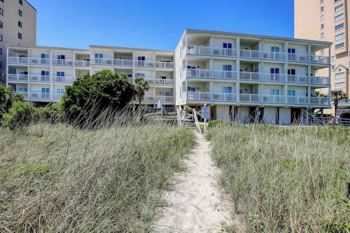 Un hotel sulla spiaggia con numerosi balconi, un sentiero sabbioso che conduce all'ingresso e un cielo azzurro terso.
