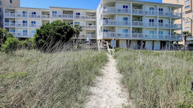 Un hôtel situé en bord de mer, doté de nombreux balcons, d’un chemin de sable menant à l’entrée et offrant une vue sur un ciel bleu azur.