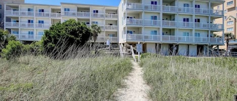 A beachfront hotel with multiple balconies, a sandy path leading to the entrance, and a clear blue sky.