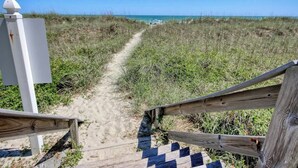 Un sentier de sable qui longe la plage et mène à l’océan, avec une clôture en bois et une structure blanche sur la gauche.