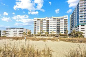 Ein Strandhotel mit mehreren Etagen, Balkonen und einem sandigen Strand im Vordergrund.
