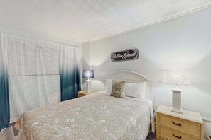 Un dormitorio con una cama, una mesita de noche, una lámpara, un cartel en la pared y una ventana con cortinas.