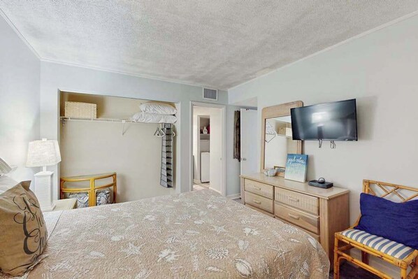 A bedroom with a bed, a television mounted on the wall, a dresser, and a chair. - Coastal Luxe | Shoreham Six C (North Myrtle Beach)