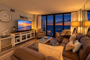 Una sala de estar con televisión, sofás, una mesa de centro y vista al océano al atardecer.