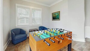 A room with a foosball table, a blue armchair, and a window with blinds. - Cloisters on the Ocean 326 (Myrtle Beach)