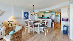 A dining area with a table and chairs, a kitchen in the background, and a living room with a sofa and lamp. - Ashworth Unit 1305 Condo (North Myrtle Beach)