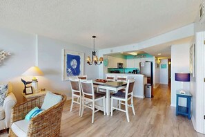 A dining area with a table and chairs, a kitchen in the background, and a living room with a sofa and lamp. - Ashworth Unit 1305 Condo (North Myrtle Beach)