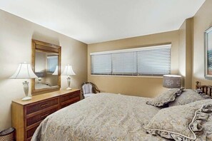 Ein Schlafzimmer mit einem Bett, zwei Nachttischlampen, einem Schminktisch, einem Sessel und einem Fenster mit Jalousien.