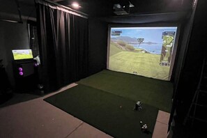A room with a large screen displaying a video game, a small table with a computer, and a green carpet. - Pawleys Paradise Retreat (Pawleys Island)