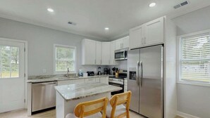 A modern kitchen with white cabinets, stainless steel appliances, and a marble island with two wooden chairs. - Pawleys Paradise Retreat (Pawleys Island)