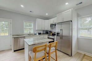 A modern kitchen with white cabinets, stainless steel appliances, and a marble island with two wooden chairs. - Pawleys Paradise Retreat (Pawleys Island)