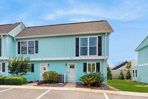 A two-story residential building with a light blue exterior, a front door, and a small front yard with landscaping. - Robbers Roost Unit 831 (North Myrtle Beach)