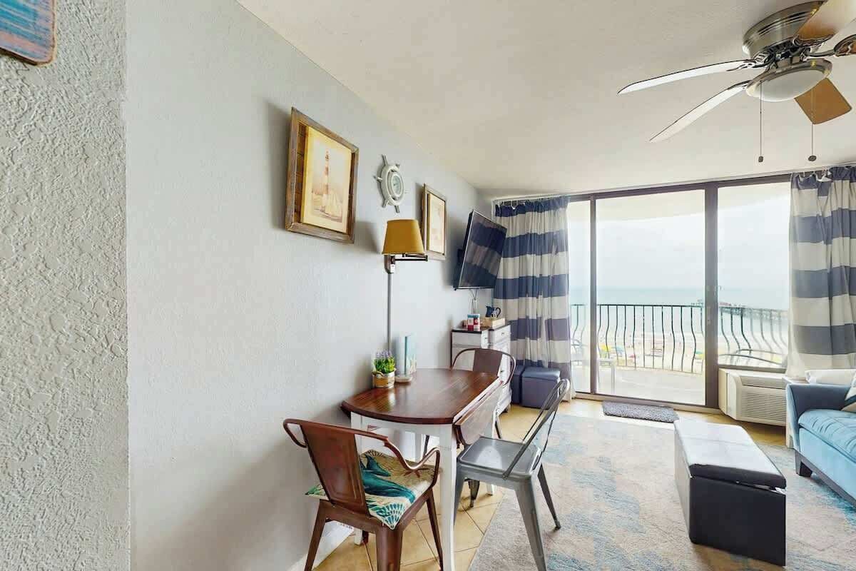 Una habitación con una mesa de comedor, sillas, un sofá, un ventilador de techo y vistas al mar a través de amplios ventanales.